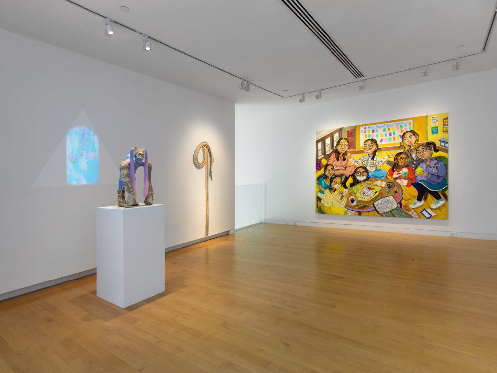 Install view of a gallery with a figurative sculpture to the left with a projection in the background, a soft sculpture hung on the wall at center and painting.