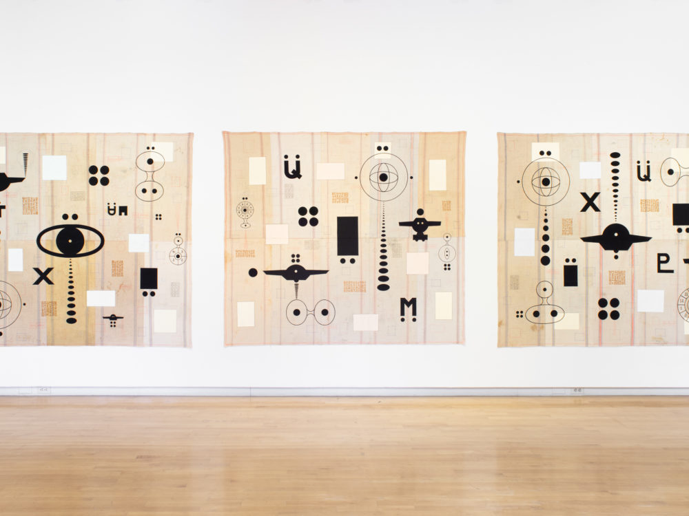 Three large-scale unstretched paintings with similar dark motifs against cream colored background hung together on a wall.