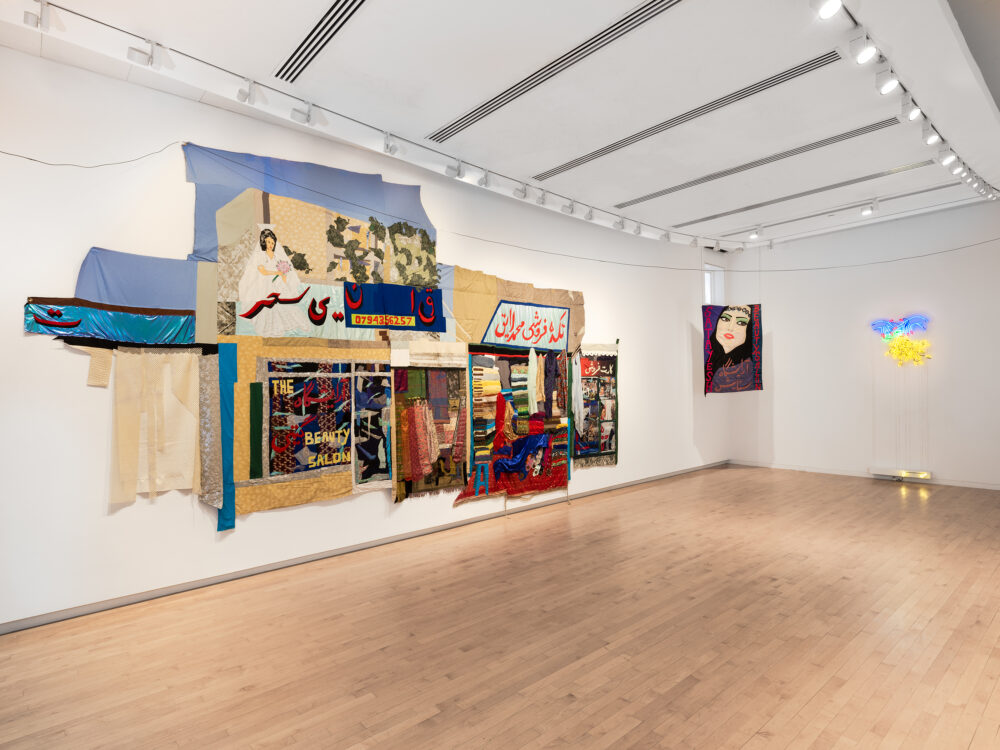 A museum gallery with large-scale tapestries hanging on the walls. A neon sign of a woman's hands with red nails and grapes hangs on the furthest wall.