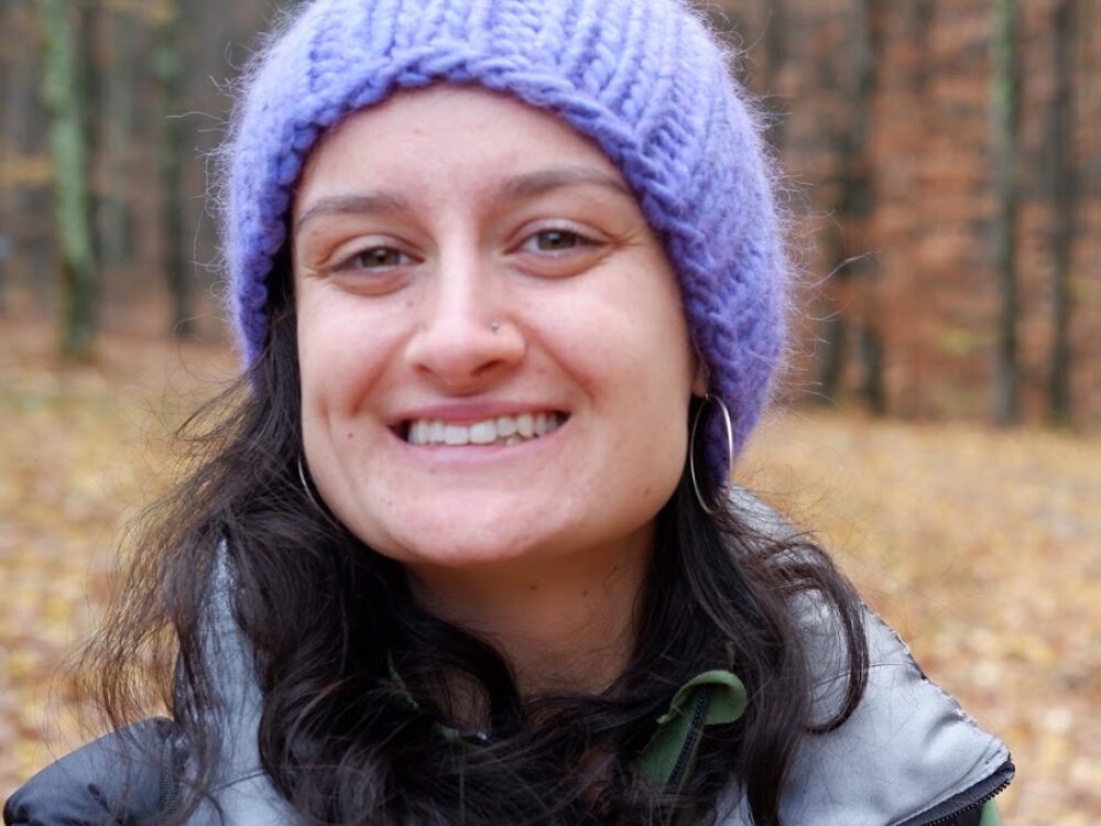 A headshot of a woman with dark hair smiling and wearing a lavender knitted hat.