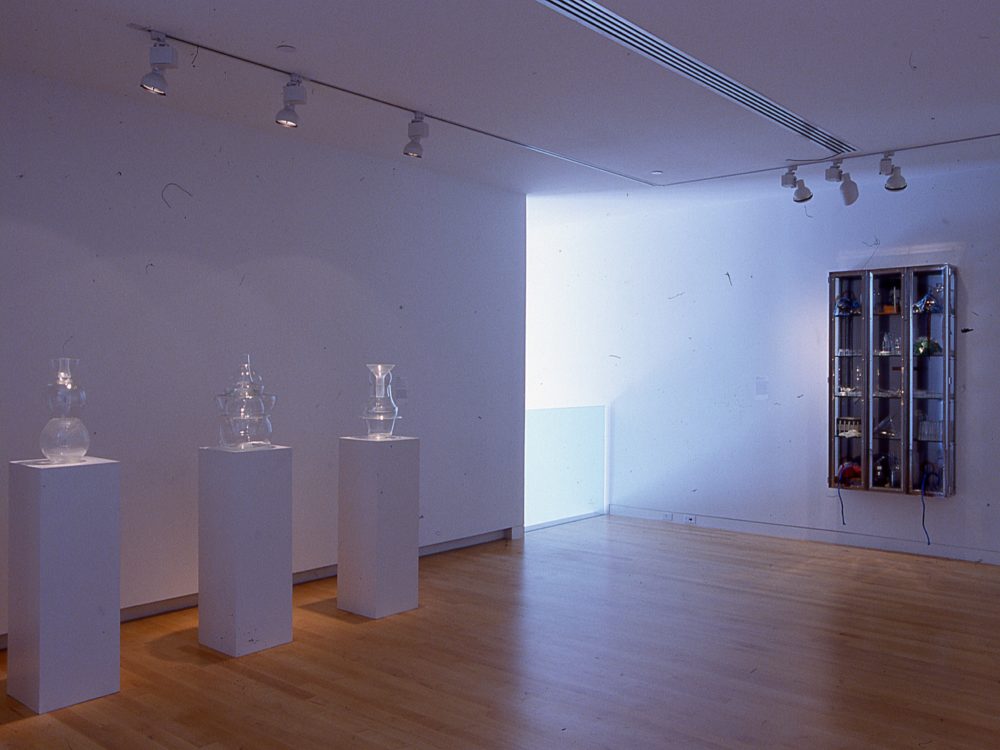 Gallery with three clear bottles on pedestals to the left; Wall mounted metal shelf with smaller bottles on right.