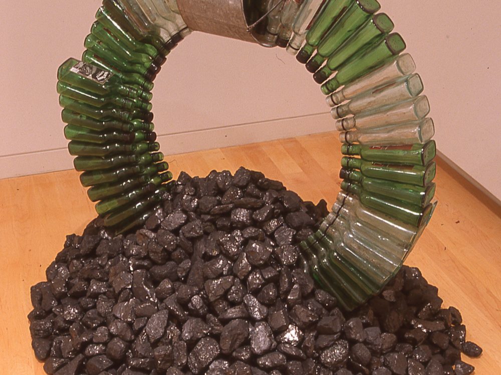 Bottles arranged in a large ring; Aluminum bucket on its side at top of ring. Ring set in a pile of black lava-like rocks that serve as base..