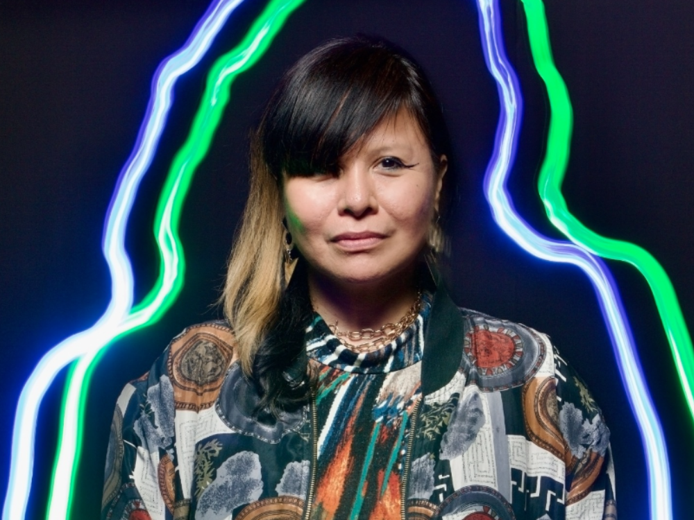 Portrait of artist, Laura Ortman, whose figure is outlined by purple and green neon in front of a dark background.